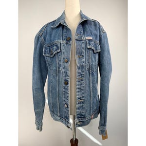 Può includere: Una giacca in denim blu a lavaggio medio con colletto classico, maniche lunghe e chiusura frontale con bottoni. La giacca presenta due tasche sul petto con patta abbottonata e bottoni in metallo. Un'etichetta rettangolare è visibile.