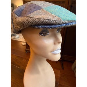 May include: A flat cap constructed from a patchwork of various wool-like fabrics in shades of brown, grey, blue, and teal. The cap features a structured brim and a multi-panel design with distinct textures and patterns across the crown and sides.