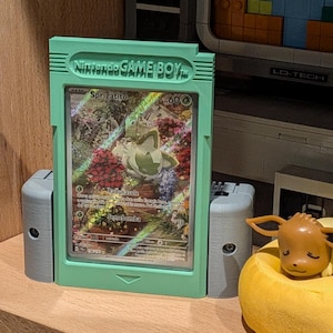 Può includere: Un porta carte verde menta in stile Game Boy con una carta Pokémon raffigurante Sprigatito. Il supporto ha due supporti laterali grigi e poggia su una mensola di legno. Una statuetta di Eevee poggia su un cuscino giallo.