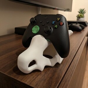 Puede incluir: Un mando de videojuegos inalámbrico negro con una cruceta verde descansa sobre un soporte blanco impreso en 3D con forma de cráneo. El mando cuenta con joysticks negros y botones A, B, X e Y de colores en el lado derecho del dispositivo.