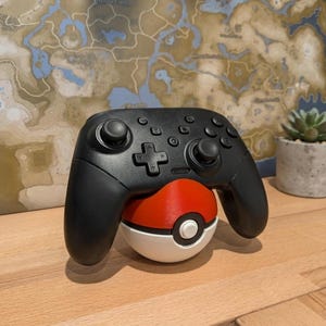 Può includere: Un controller wireless nero per videogiochi appoggiato su un supporto sferico rosso e bianco progettato per assomigliare a una Pokeball. Il controller presenta due levette analogiche, un pad direzionale e vari pulsanti di azione sulla parte anteriore.