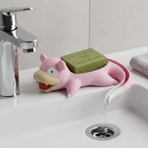May include: A pink Slowpoke-shaped soap dish holding a green bar of soap. The dish has a white and pink tail that directs water into the sink. The soap is labelled "NATURAL OLIVE OIL SOAP". The sink has a chrome tap.