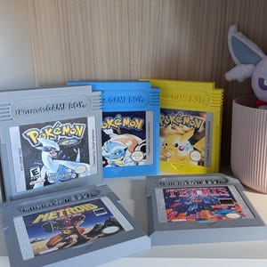 Op de afbeelding: Vijf Nintendo Game Boy-cartridges waaronder Pokémon Silver, Blue en Yellow versies, plus Metroid en Tetris. Een klein paars pluche wezen zit in een lichtroze geribbelde pot achter de gele Pokémon Yellow-gamecartridge.