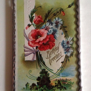 May include: A vintage birthday greeting card with a silver frame. The card features a floral design with red poppies, blue flowers, and green leaves. The text "Birthday Greeting" is written in a decorative script.