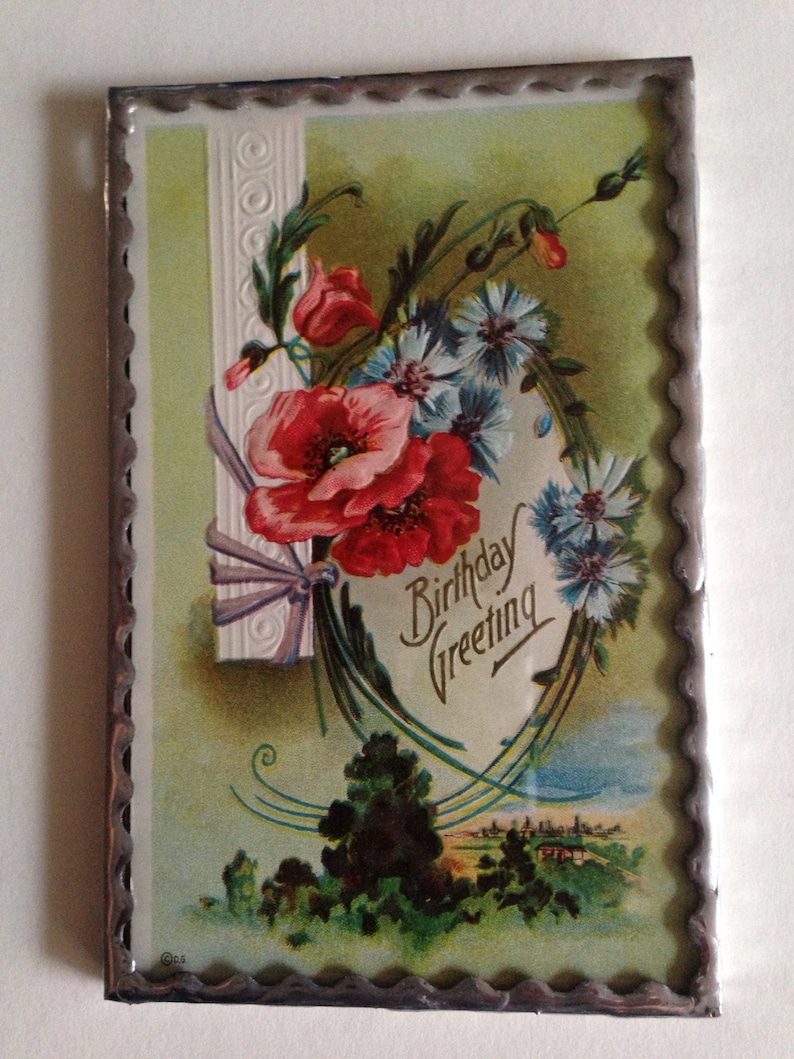 May include: A vintage birthday greeting card with a silver frame. The card features a floral design with red poppies, blue flowers, and green leaves. The text "Birthday Greeting" is written in a decorative script.