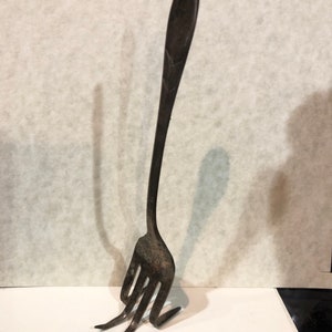May include: A dark, weathered metal fork with a long, slender handle and three tines that are bent outward at the base, allowing the utensil to stand upright on its own. The surface of the metal shows signs of oxidation and a matte, textured finish.