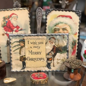 May include: A rectangular Christmas sign with a scalloped metal border featuring a vintage illustration of a child and Santa Claus with the text I wish you a very MERRY CHRISTMAS, mounted on a silver spoon handle stand.