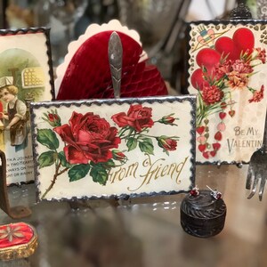 May include: Four vintage Valentine's Day cards with red hearts and floral designs. The cards are displayed on a table with a small, round, wooden box and a silver fork.
