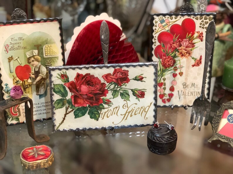 May include: Four vintage Valentine's Day cards with red hearts and floral designs. The cards are displayed on a table with a small, round, wooden box and a silver fork.