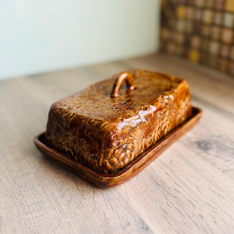 May include: A rectangular brown ceramic butter dish with a glossy finish and a textured floral pattern embossed across the lid. The lid features a small arched handle on top, and the base is a matching rectangular tray with raised edges.