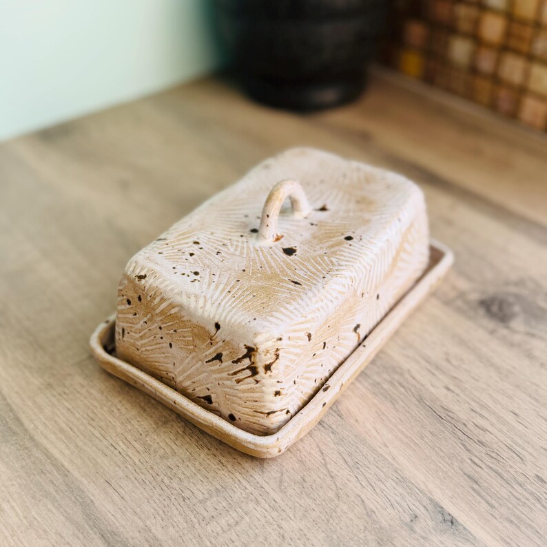 May include: A rectangular ceramic butter dish with a lid featuring a small arched handle. The surface is off-white with a textured, carved sunburst pattern and dark brown speckles throughout the exterior of the lid and the base tray.