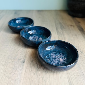 May include: A three-sectioned ceramic serving dish. The dish is a deep blue with a speckled, glazed finish. The three connected bowls are oval-shaped and sit on a light-coloured wooden surface. The dish is ideal for serving snacks or dips.