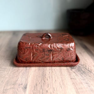 May include: A rectangular ceramic butter dish with a lid featuring a dark reddish-brown glaze and a textured, leaf-like pattern. The lid has a small, arched handle on top, and the base is a matching flat tray with raised edges.