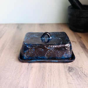 May include: A rectangular ceramic butter dish with a dark blue and black glaze. The lid features a repeating embossed fan-like pattern and a small arched handle on top. The base is a matching rectangular tray with slightly raised edges.