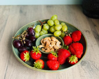 May include: A round, sage green ceramic serving dish with a central bowl containing roasted cashews. The outer rim holds a cluster of dark purple grapes, a bunch of light green grapes, and several bright red strawberries with green stems.