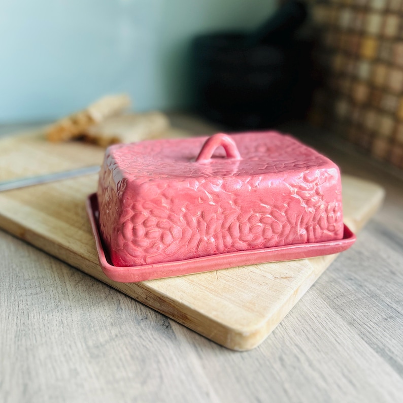 Handmade ceramic butter dish with lid, embossed flower pattern, rose pink glaze, placed on a wooden board