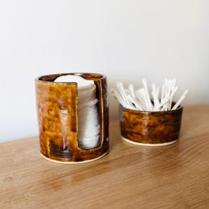 May include: A cylindrical ceramic container with a glossy brown glaze and a vertical cutout, filled with a stack of white cotton rounds, positioned next to a matching shallow ceramic bowl containing a cluster of white cotton swabs.