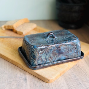 May include: A blue ceramic butter dish with a lid and handle, featuring a textured, fan-like pattern. The dish sits on a wooden chopping board, with slices of bread in the background.