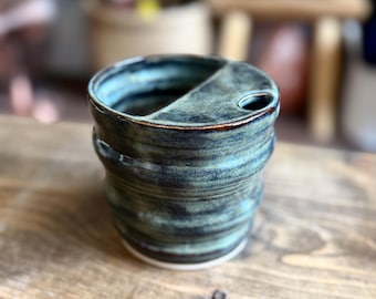 May include: A ceramic travel mug with a dark teal and blue glaze featuring a built-in lid with a small circular opening. The mug has a ribbed, cylindrical body with a smooth finish and a white base, showcasing a deep, mottled color palette.