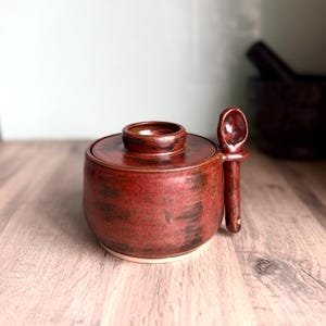 May include: A round, dark red ceramic sugar bowl with a matching lid and a small spoon resting in a side holder. The glaze features mottled dark brown and black accents across the surface, creating a textured appearance on the clay vessel.