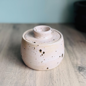 May include: A round ceramic lidded jar in a pale pink hue with a speckled finish featuring dark brown spots. The container has a smooth, curved body and a matching lid with a small, raised central knob handle on top for easy removal.