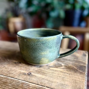 May include: A ceramic mug with a wide, rounded bowl and a single handle. The exterior features a mottled glaze in shades of forest green, olive, and hints of yellow, creating a textured, earthy appearance with a smooth, glossy finish throughout.