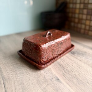 May include: A rectangular ceramic butter dish with a deep reddish-brown glaze and a textured, repeating leaf-like pattern embossed across the lid. The lid features a small, arched handle on top, and it rests on a matching flat base.