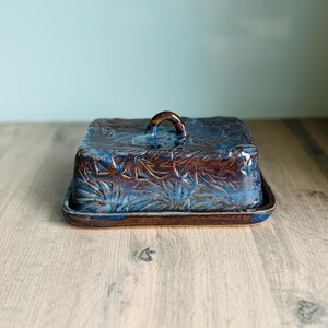 May include: A rectangular ceramic butter dish with a lid featuring a small arched handle. The piece has a deep brown and vibrant blue glaze with a textured, leaf-like pattern embossed across the entire surface of the lid and base.