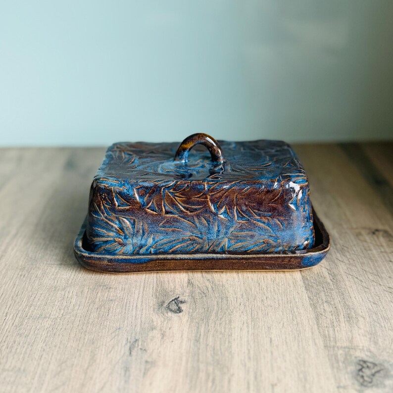 May include: A rectangular ceramic butter dish with a lid featuring a small arched handle. The piece has a deep brown and vibrant blue glaze with a textured, leaf-like pattern embossed across the entire surface of the lid and base.