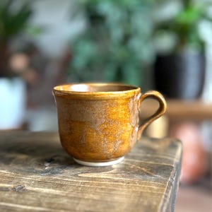 May include: A handmade ceramic mug with a textured, golden-brown glaze. The mug has a rounded body, a small white base, and a curved handle. The mug is sitting on a wooden surface.