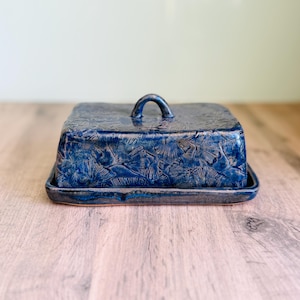 May include: A rectangular ceramic butter dish with a deep blue glaze and a textured ginkgo leaf pattern. The lid features a small arched handle on top, and the base has a matching blue finish with a slightly raised edge for the lid to rest.