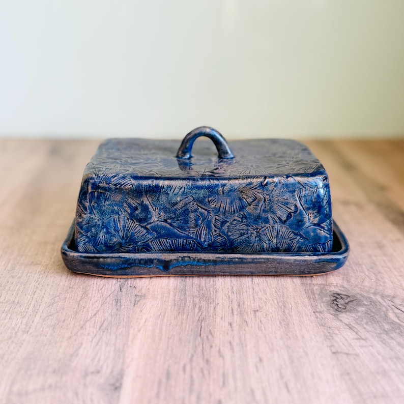 May include: A rectangular ceramic butter dish with a deep blue glaze and a textured ginkgo leaf pattern. The lid features a small arched handle on top, and the base has a matching blue finish with a slightly raised edge for the lid to rest.