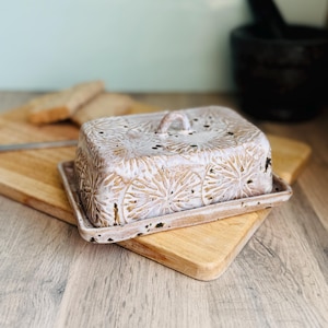 May include: A ceramic butter dish with a lid, decorated with a floral pattern in white and beige. The dish sits on a matching tray, with a small handle on top. The butter dish is on a wooden chopping board.