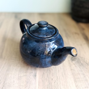 May include: A dark blue ceramic teapot with a glossy finish. The teapot features a rounded shape, a curved spout, and a loop handle. The lid is topped with a small knob. It sits on a light wooden surface.