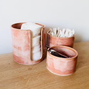 May include: Three cylindrical ceramic containers with a mottled pink and peach glaze. The tallest container features a vertical cutout holding a stack of white cotton rounds. A medium container holds white cotton swabs, and a small bowl holds tweezers.