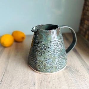 May include: A ceramic jug with a tapered body and a curved handle. The exterior features a mottled teal and green glaze with a repeating embossed floral pattern. The rim has a small pouring spout, and the base shows a light-coloured clay rim.