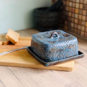 May include: A blue ceramic butter dish with a textured pattern and a small handle on the lid. The dish sits on a matching base. The butter dish is on a wooden chopping board with a knife and two slices of bread.