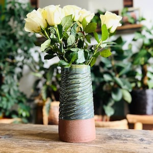 May include: A ceramic vase with a textured, dark green glazed upper section and a smooth, unglazed terracotta base. The vase is filled with a bouquet of pale yellow roses with green leaves, creating a contrast between the floral and ceramic.