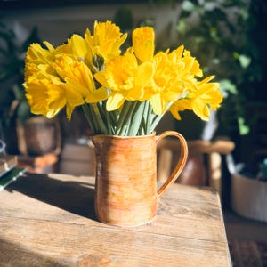 May include: A collection of vibrant yellow daffodils in a rustic, orange ceramic jug. The jug has a curved handle and is placed on a wooden surface, with sunlight highlighting the flowers. The background is blurred.