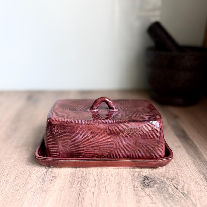 May include: A rectangular ceramic butter dish with a deep burgundy glaze and a textured, fan-like pattern across the surface. The lid features a small, arched handle at the top, and the base has a matching raised rim for secure placement.