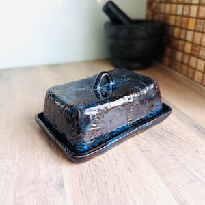 May include: A rectangular ceramic butter dish with a dark blue and black glazed finish. The lid features a textured, repeating floral pattern and a small arched handle on top. The base is a matching flat tray with raised edges.