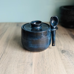May include: A round, dark blue ceramic sugar bowl with a matching lid and a small, integrated side loop holding a coordinating ceramic spoon. The glaze features deep navy and black tones with a glossy finish and a smooth, cylindrical body shape.