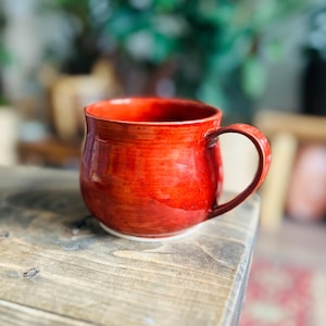 May include: A handmade, red ceramic mug with a rounded body and a curved handle. The mug has a glossy finish and sits on a white base. The mug is designed for hot beverages.