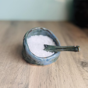 May include: A small, round, handmade ceramic bowl with a mottled blue and green glaze, filled with white granular salt. A matching ceramic spoon with a dark, glazed finish rests inside the bowl, partially submerged in the white salt crystals.