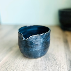 May include: A dark blue ceramic creamer with a silver rim and a small spout. The creamer has a textured surface. The background features a light blue wall and a wooden surface.