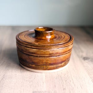 May include: A brown glazed ceramic container with a lid. The lid has a small handle and concentric circles. The container sits on a white base. The background is a neutral colour.