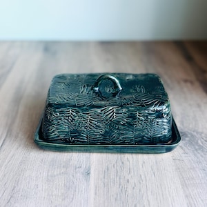 May include: A rectangular ceramic butter dish with a dark teal, glossy glaze. The lid features a textured, imprinted pattern of overlapping fern leaves and a small, arched handle on top. The base is a matching flat, rectangular tray.