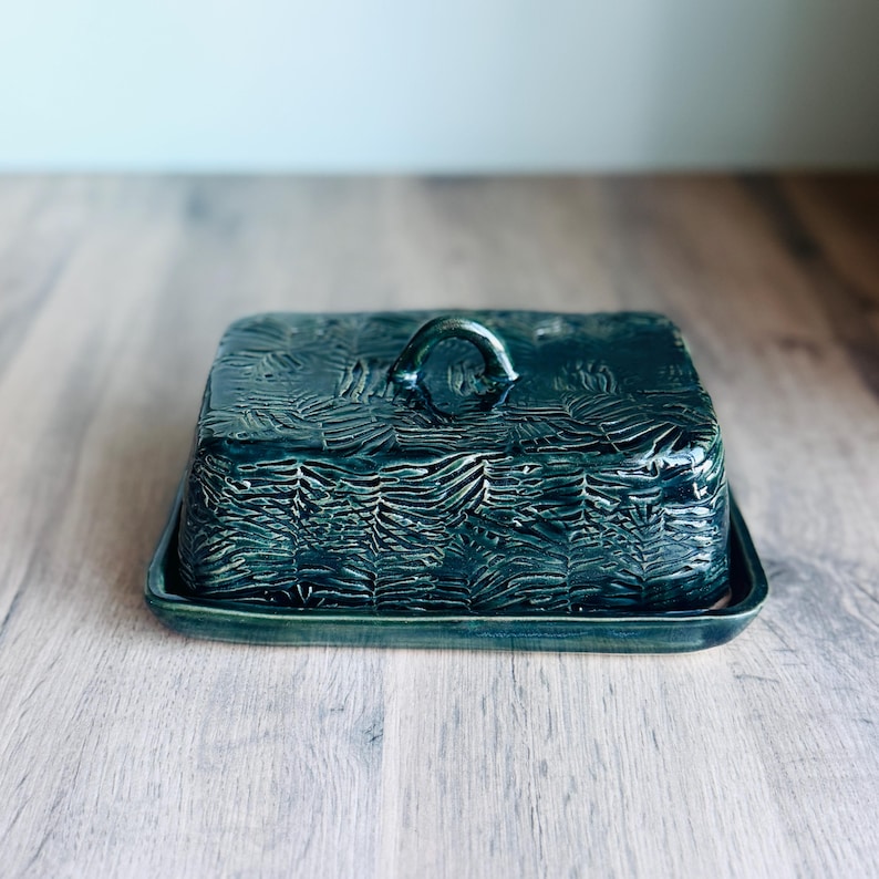 May include: A rectangular ceramic butter dish with a dark teal, glossy glaze. The lid features a textured, imprinted pattern of overlapping fern leaves and a small, arched handle on top. The base is a matching flat, rectangular tray.