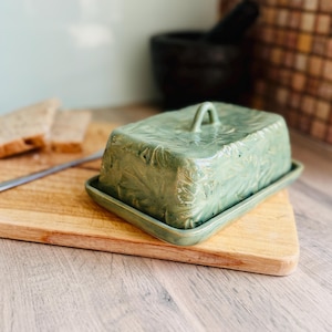 May include: A rectangular ceramic butter dish with a sage green glaze and a textured leaf pattern embossed on the lid. The dish features a matching base tray and a small arched handle on top of the lid, providing a functional kitchen storage piece.