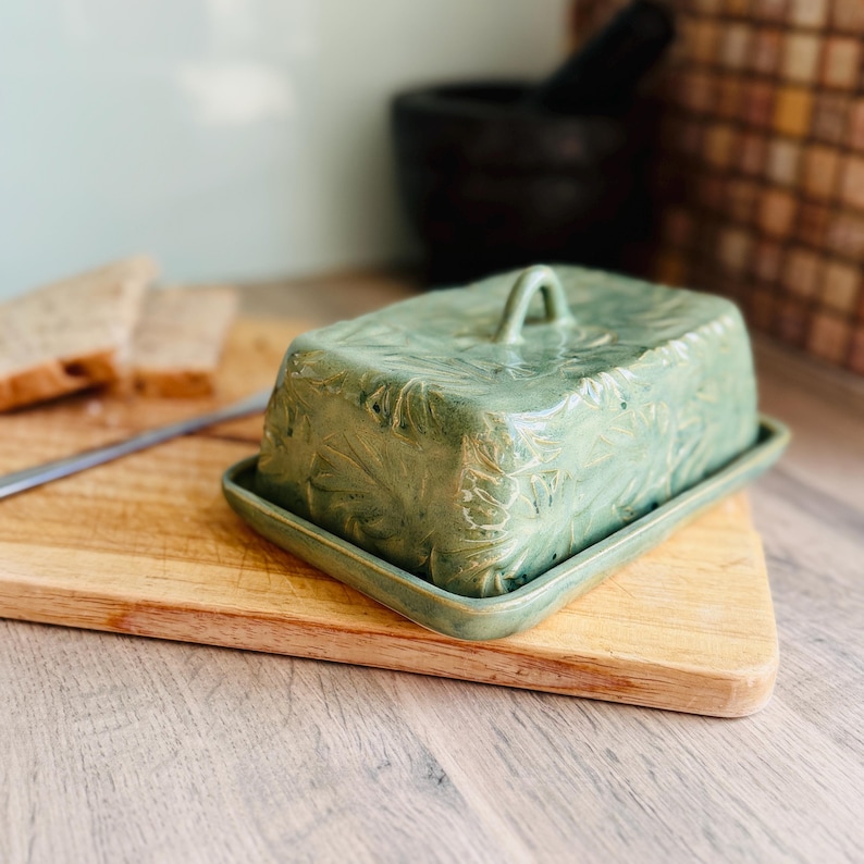 May include: A rectangular ceramic butter dish with a sage green glaze and a textured leaf pattern embossed on the lid. The dish features a matching base tray and a small arched handle on top of the lid, providing a functional kitchen storage piece.